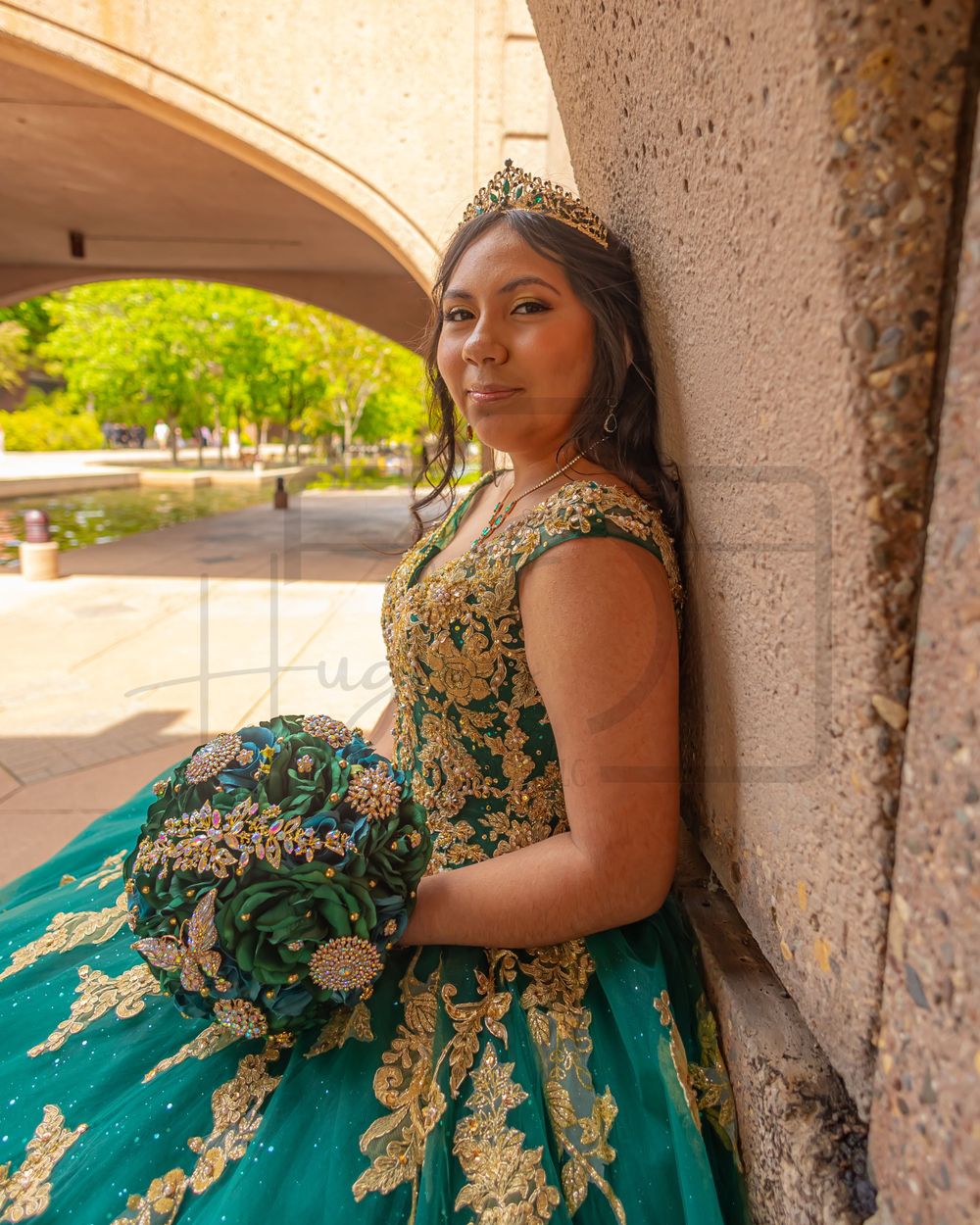 A regal portrait in an emerald green and gold quinceañera gown against a stone archway.