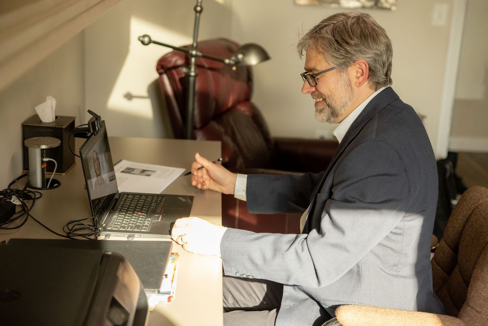 Professional man in suit working on laptop at desk in home office, engaged in video conference meeting.