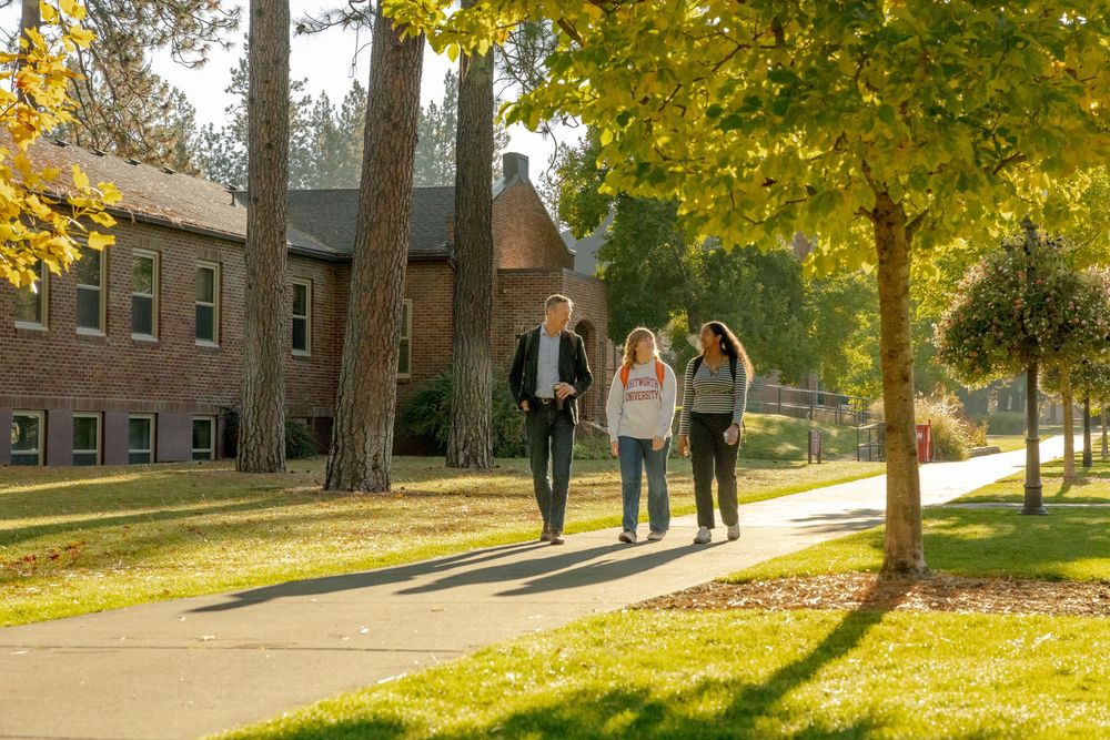 Three students walk along a tree-lined campus path beside a brick building on a sunny autumn day.