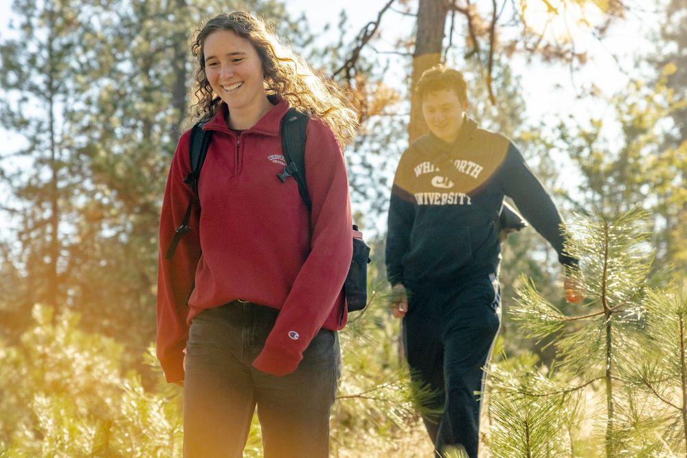 Two students hiking through a sunlit forest trail, wearing university sweatshirts and backpacks, smiling outdoors.