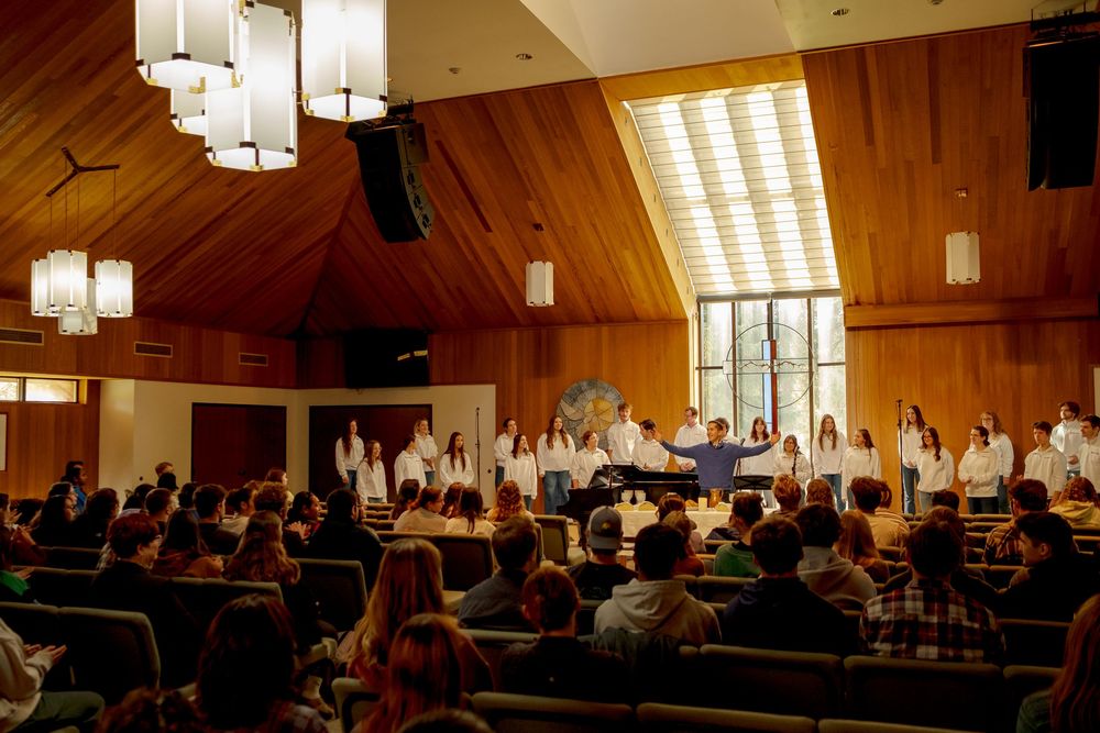 A choir performs in a wood-paneled chapel with pendant lights while an audience watches attentively.