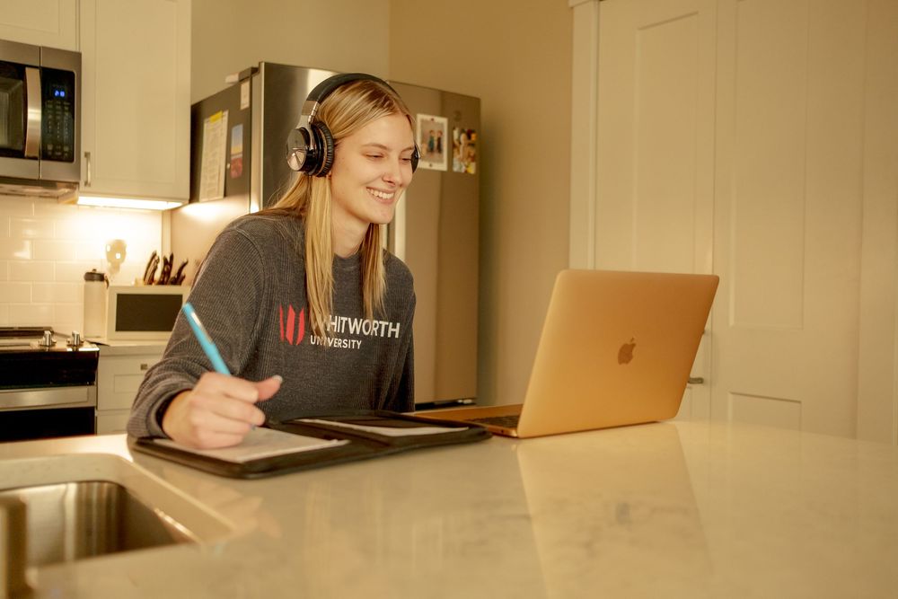 Whitworth University student studying online with laptop and headphones at kitchen counter while taking notes.