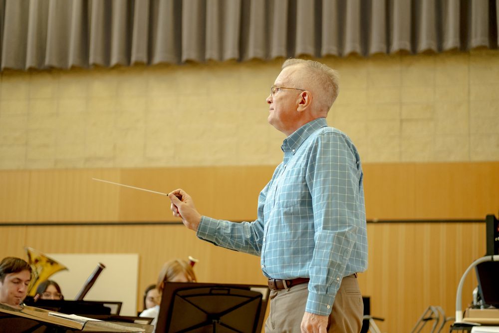 An orchestra conductor leads a rehearsal, holding a baton in a blue plaid shirt with musicians and music stands visible.