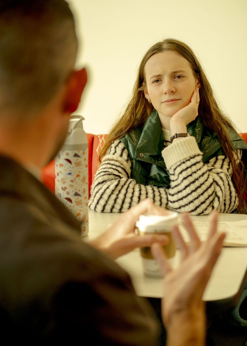 Young woman in striped sweater and green scarf listens attentively during a conversation at a table.