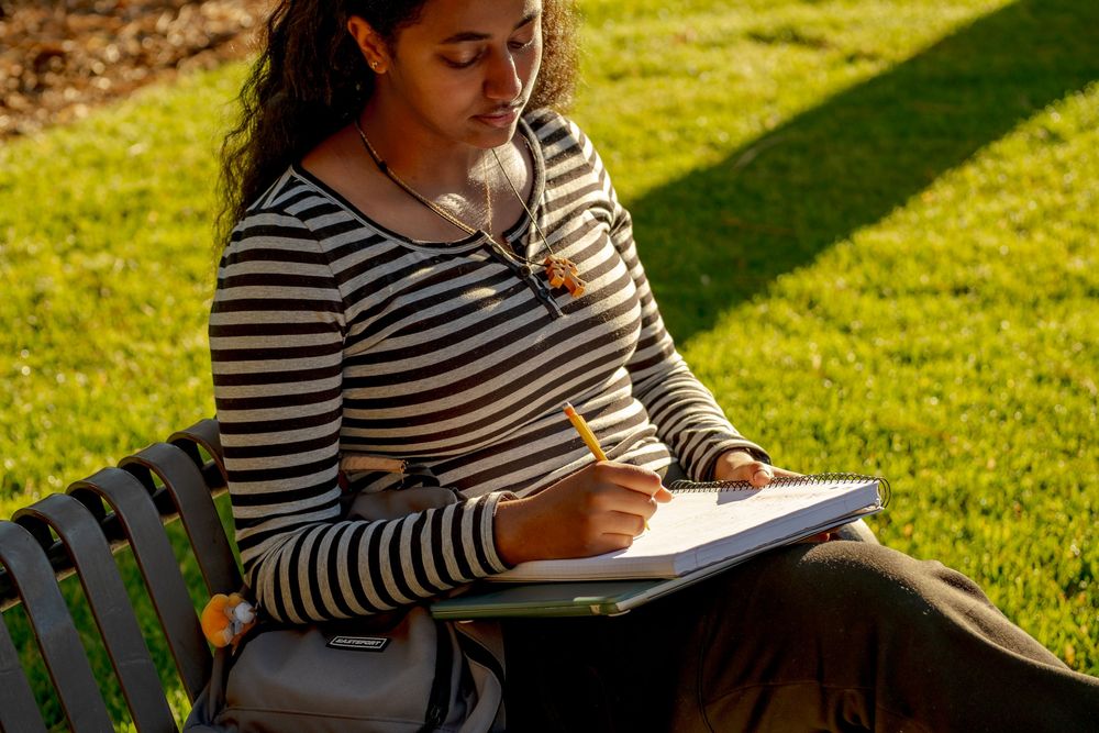 A young woman writes in a notebook with a pencil while sitting on a bench outdoors in sunlight.