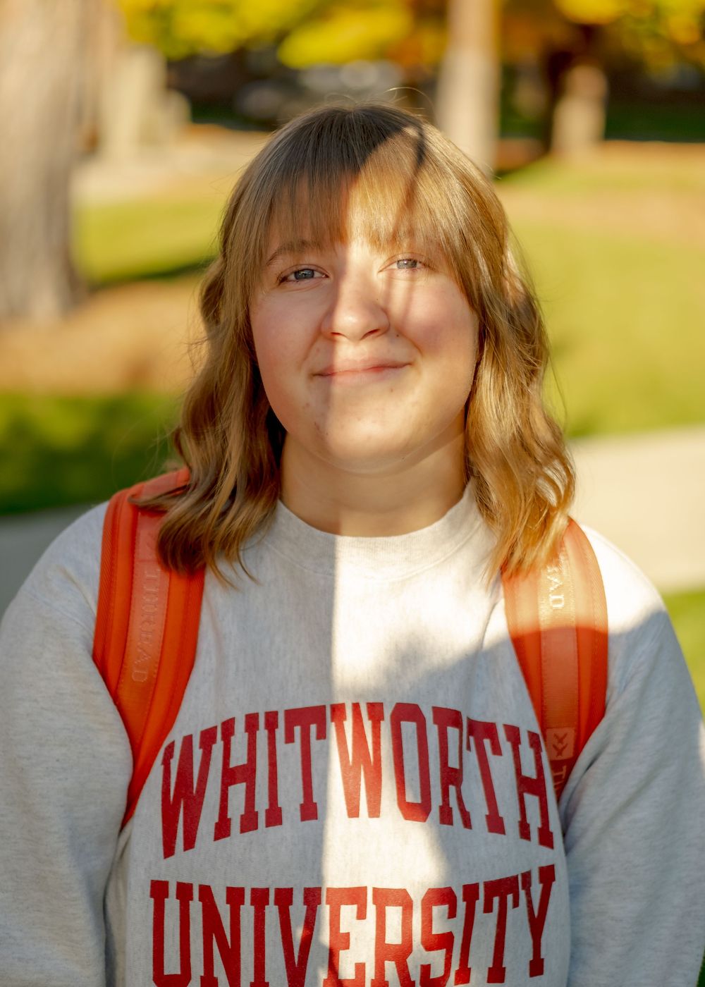 Smiling Whitworth University student wearing a gray sweatshirt and red backpack on a sunny campus.