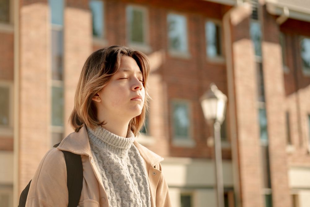 Young woman with eyes closed enjoying sunlight outdoors near brick building on sunny day.
