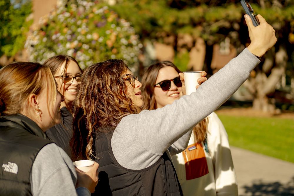 Four young women smiling and taking a selfie outdoors on a sunny day, holding coffee cups on a college campus.