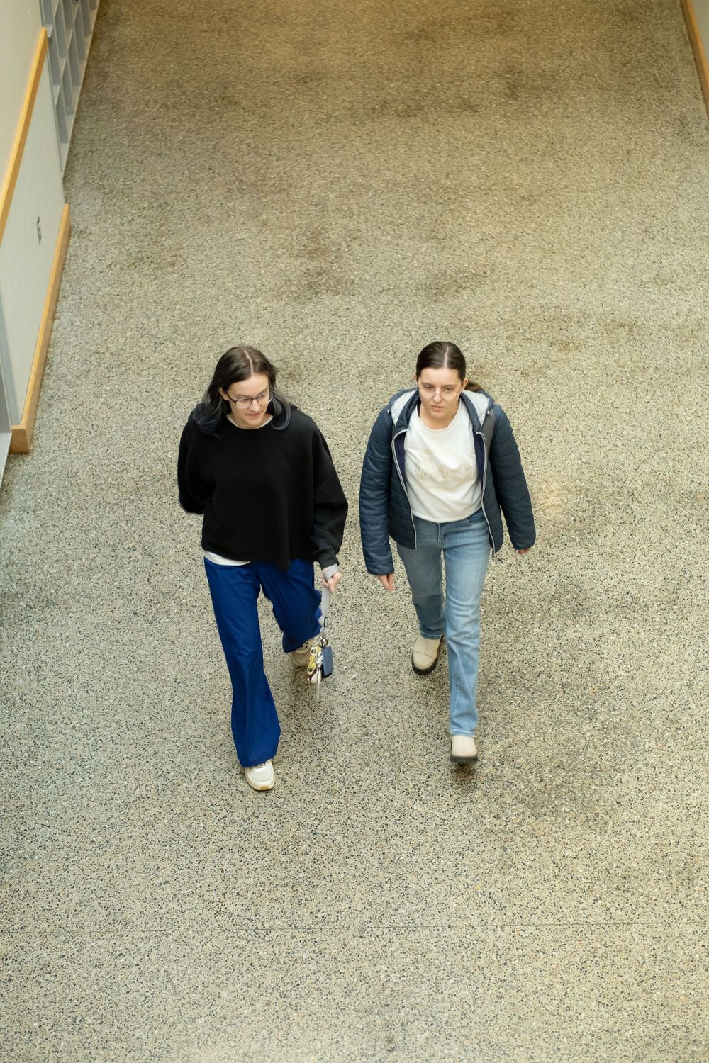 Two young women walking together down a hallway, one in a black hoodie and one in a blue jacket, both wearing jeans.