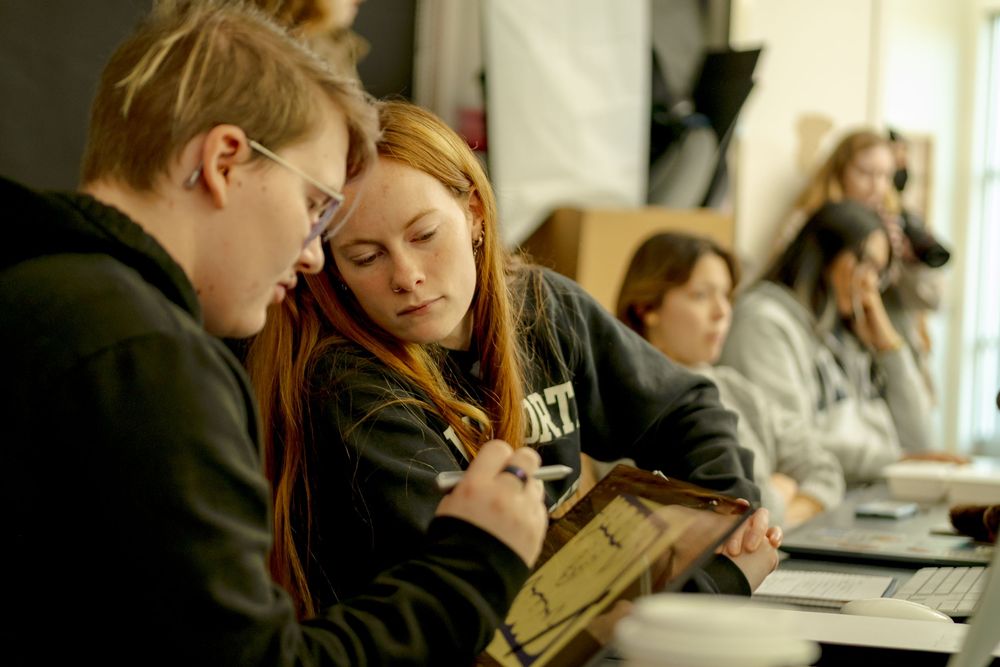 Two female students collaborate on notes together in a classroom setting with peers working in the background.