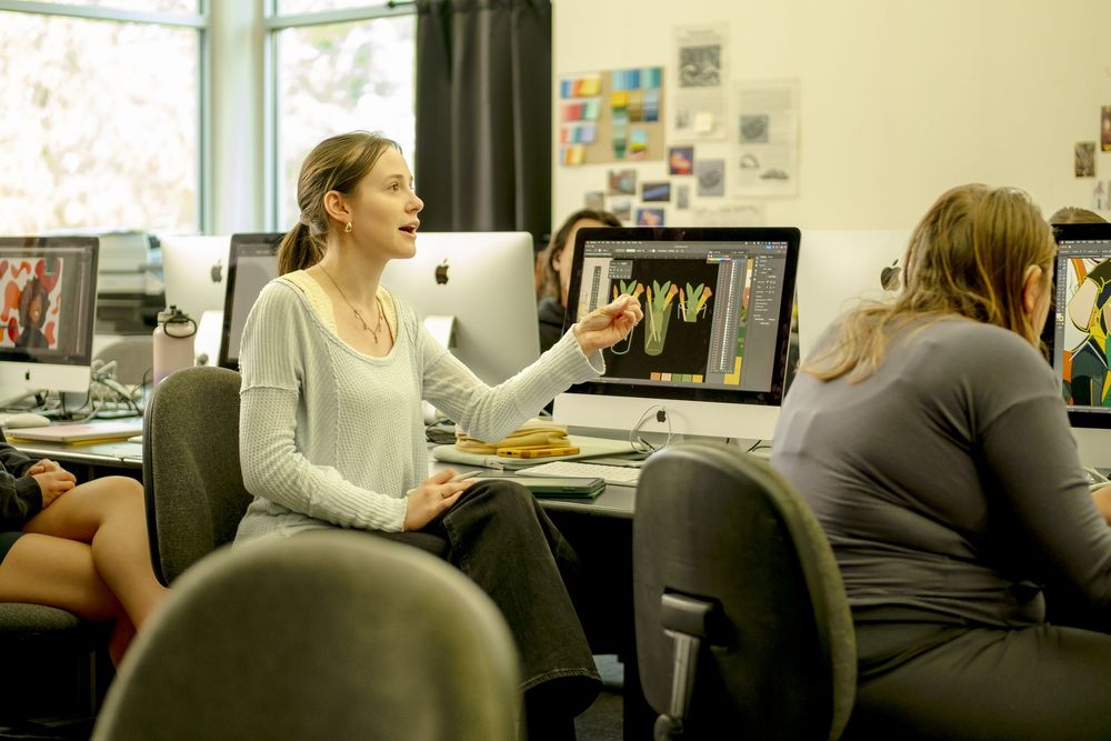 A woman in a white sweater gestures toward an iMac screen in a busy graphic design classroom.