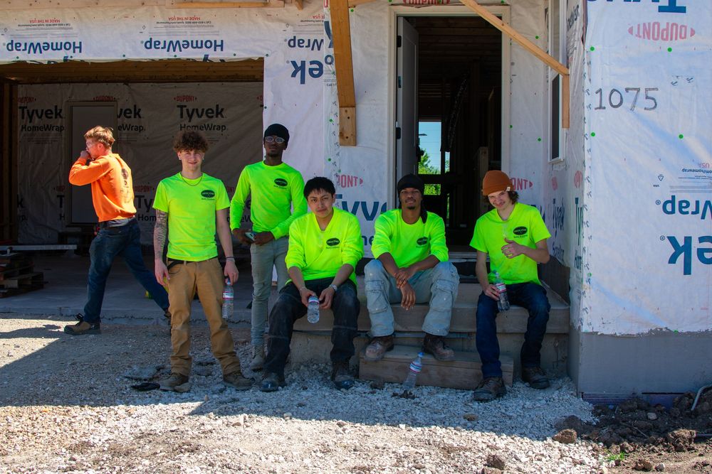 Six construction workers in neon green shirts pose outside a building under construction wrapped in Tyvek house wrap.