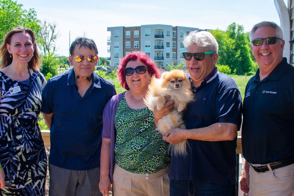 Group of five adults smiling outdoors holding a Pomeranian dog, with a modern apartment building in the background.
