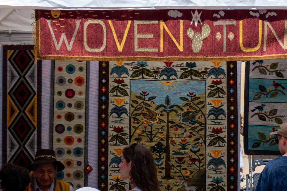 Colorful woven tapestries and rugs displayed at a Woven Tun market stall with visitors browsing.