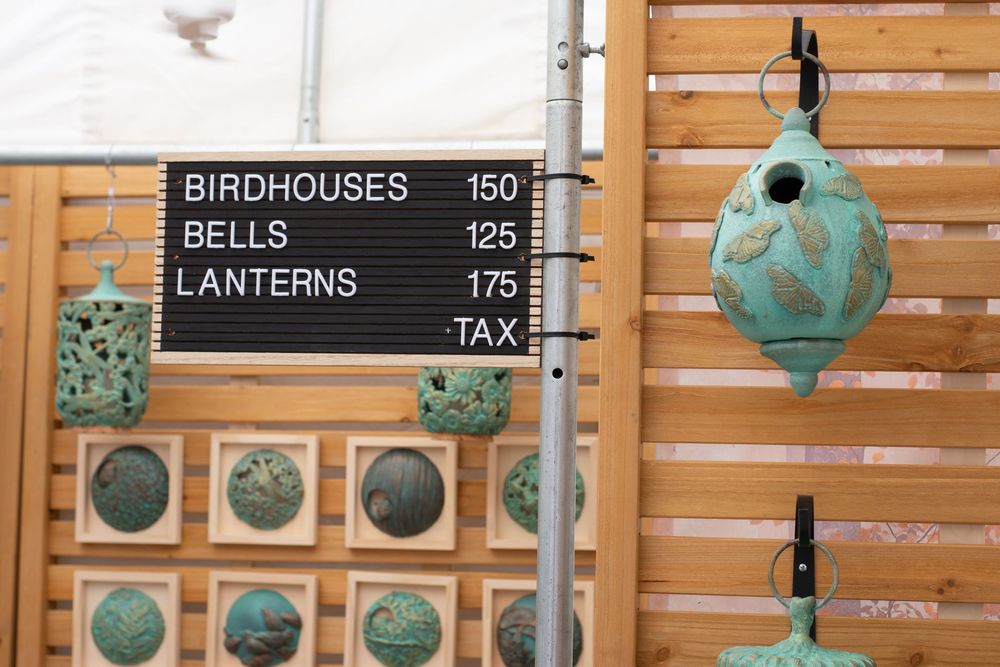 Handcrafted turquoise patina birdhouses, bells, and lanterns displayed at an outdoor market with a price sign.