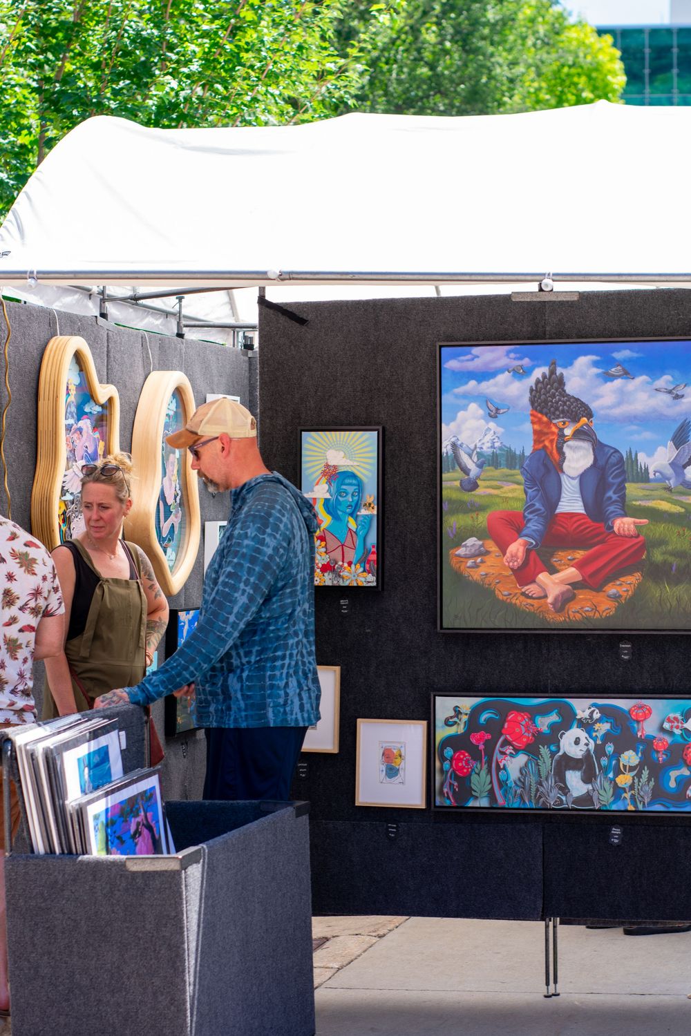 People browsing colorful paintings at an outdoor art fair booth under a white canopy on a sunny day.