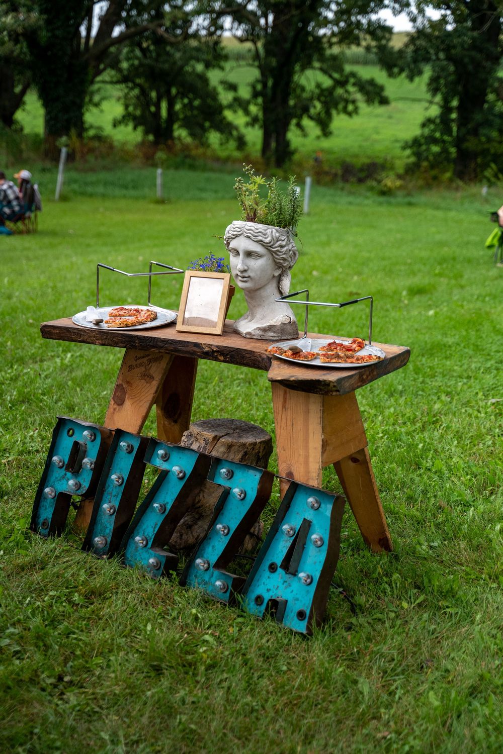 Outdoor pizza station with marquee letter signs, rustic wooden table, classical bust planter, and food displays on green lawn.