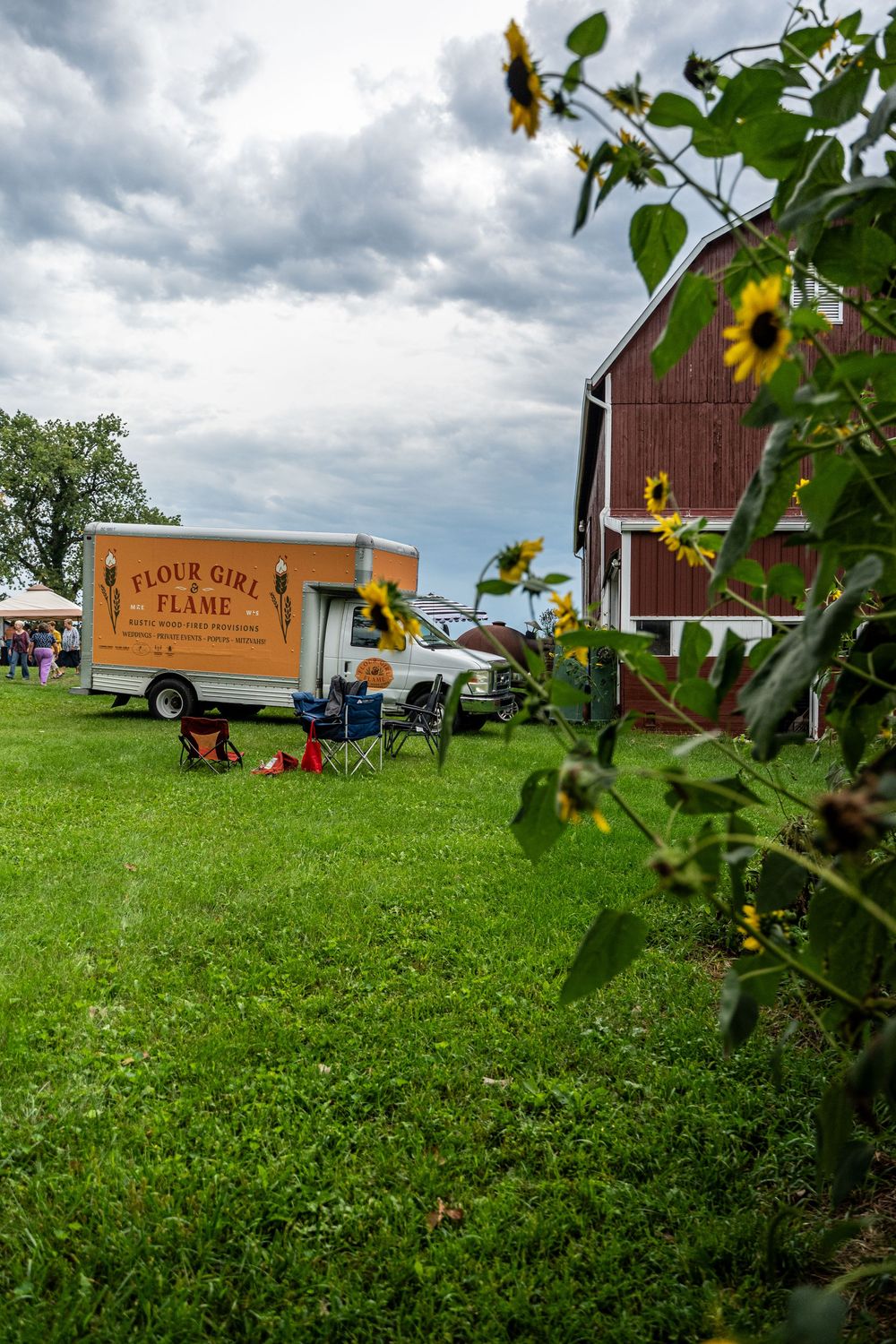 Flour Girl & Flame food trailer parked on green grass beside a red barn with sunflowers under cloudy skies.