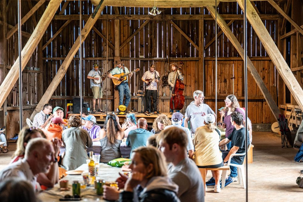 Live bluegrass band performs on stage inside a rustic wooden barn while guests dine at tables during a farm event.