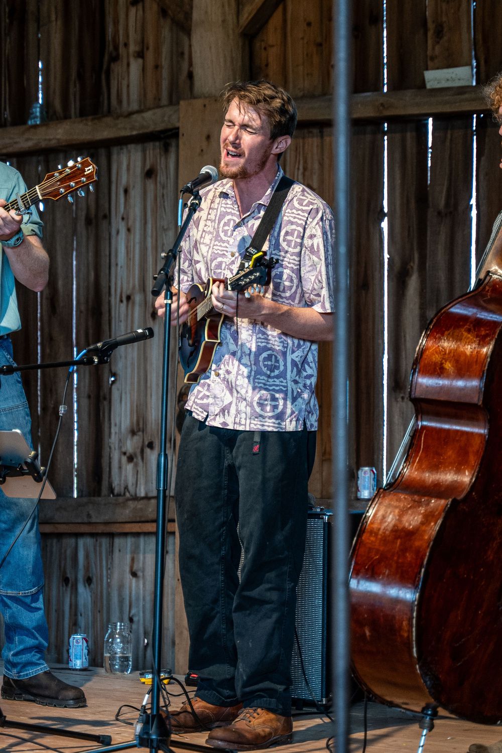 Man playing guitar and singing into microphone on rustic barn stage with upright bass visible nearby.
