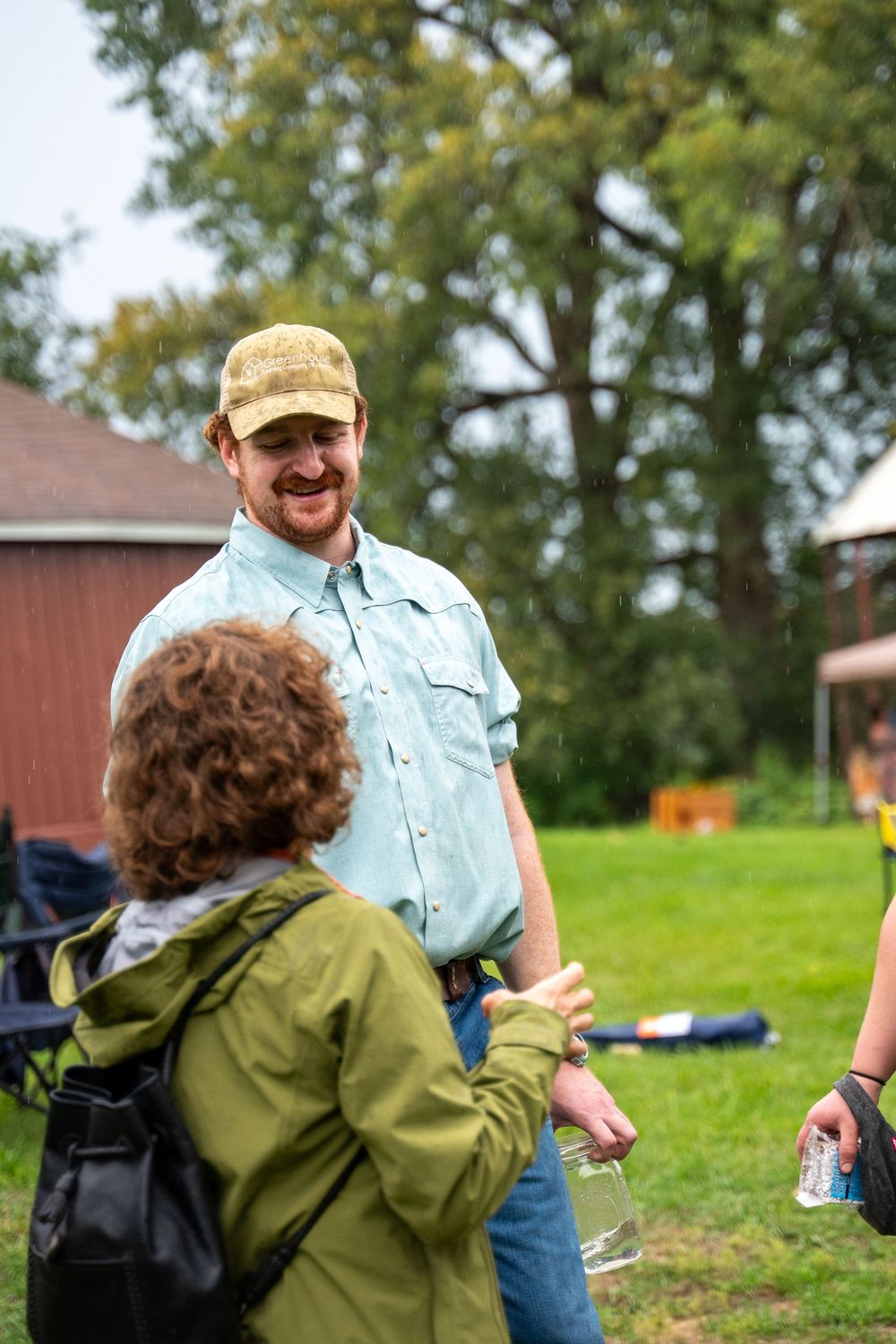 Man in cap and blue shirt smiling outdoors at a rural gathering near a red barn.