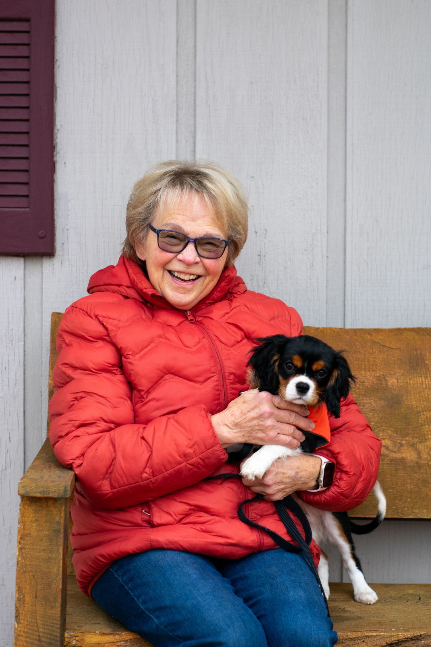 A smiling senior woman in a red puffer jacket holds a small Cavalier King Charles Spaniel puppy.