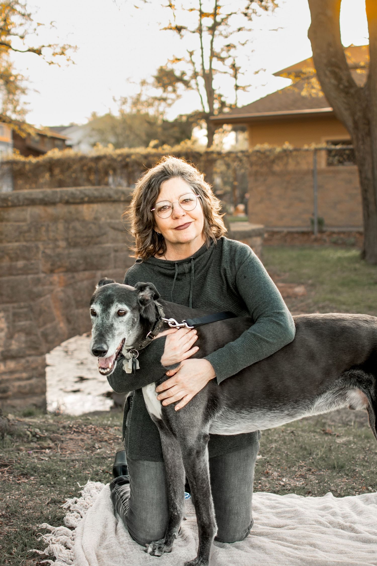 Smiling woman with glasses hugging a senior grey greyhound dog outdoors in a backyard setting.