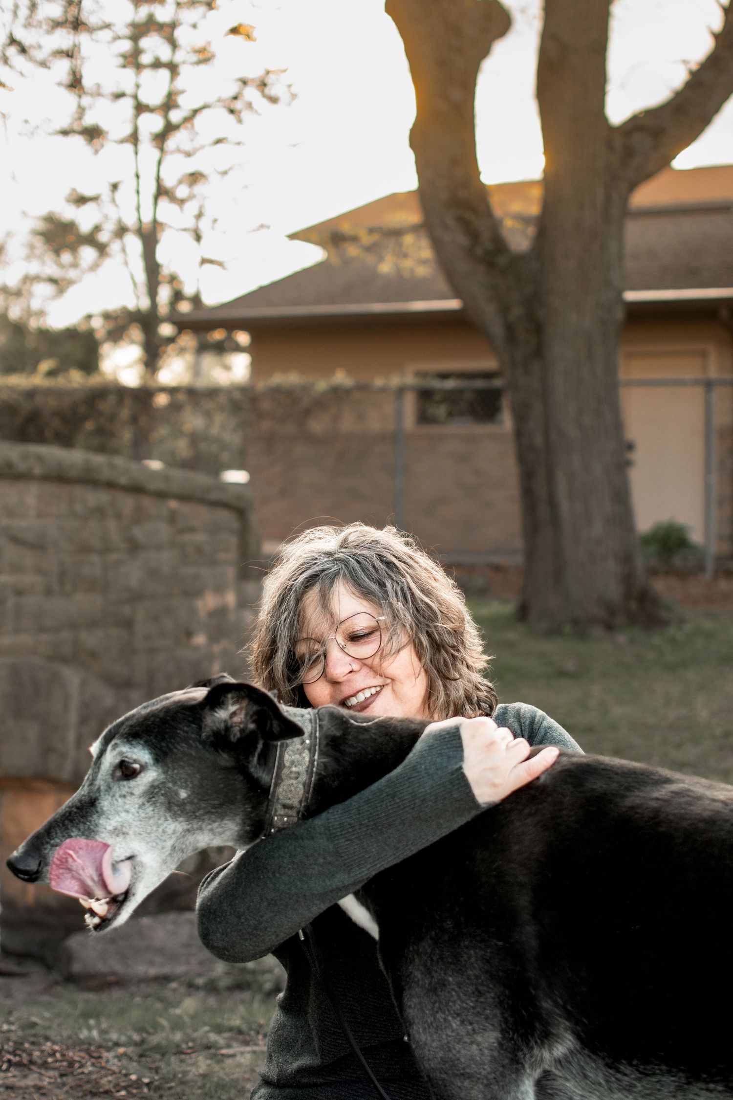 A woman with curly hair and glasses hugs a large black senior dog in a backyard.