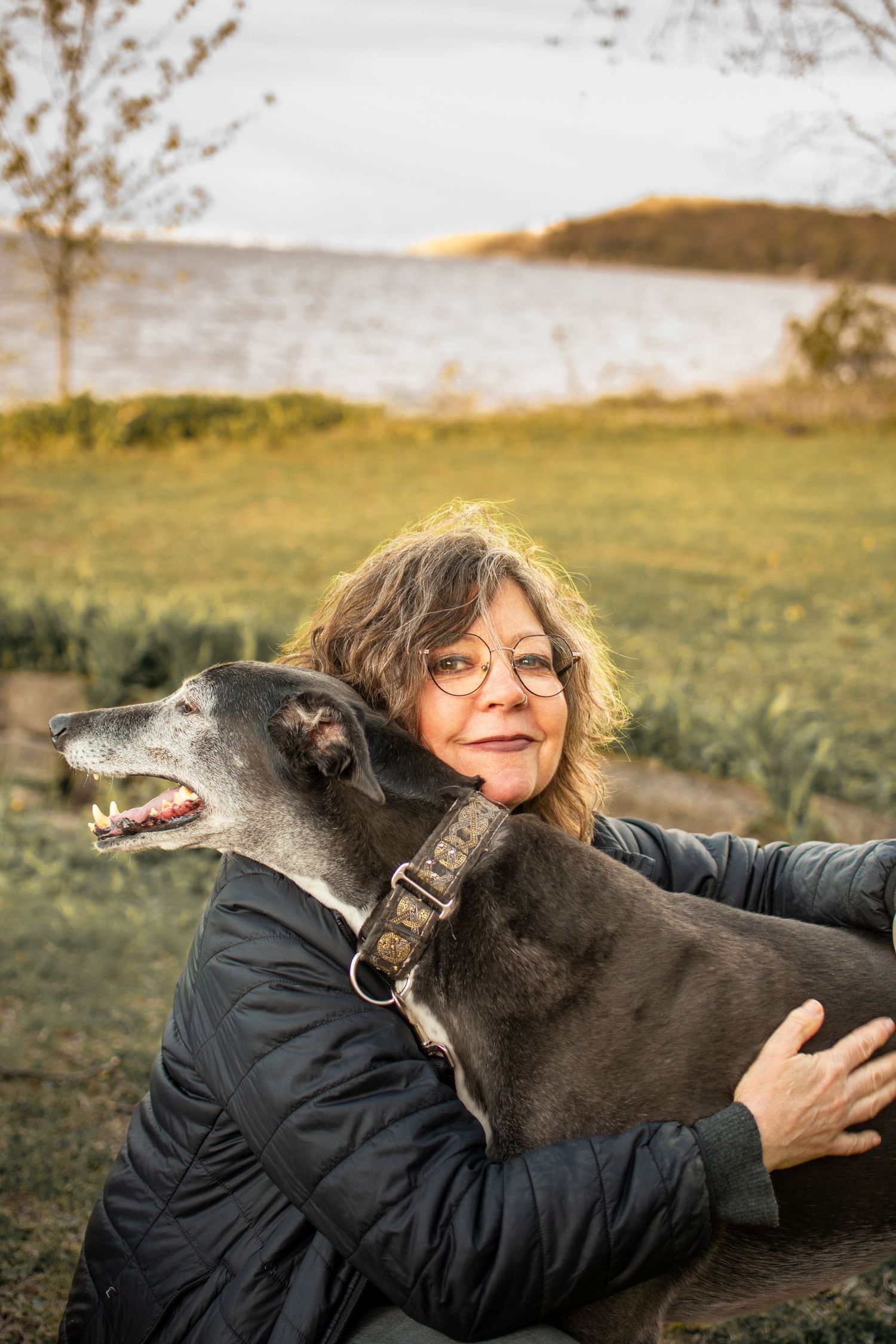 A woman with curly hair and glasses embraces a large black dog near a scenic waterfront.