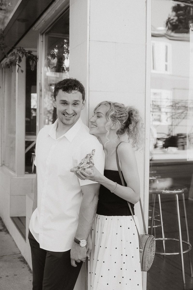 Black and white photo of a smiling couple posing outside a cafe, woman in polka dot skirt whispering to man in white shirt.