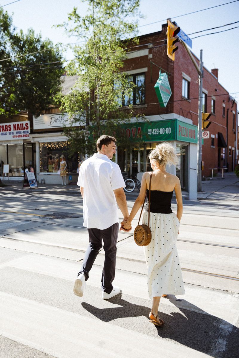 Couple holding hands crossing a sunny urban street, woman in black top and white skirt, man in white shirt.