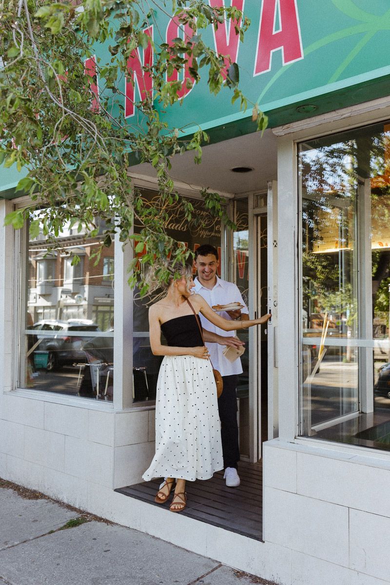 Couple smiling and holding food outside a green Nova restaurant storefront on a sunny day.