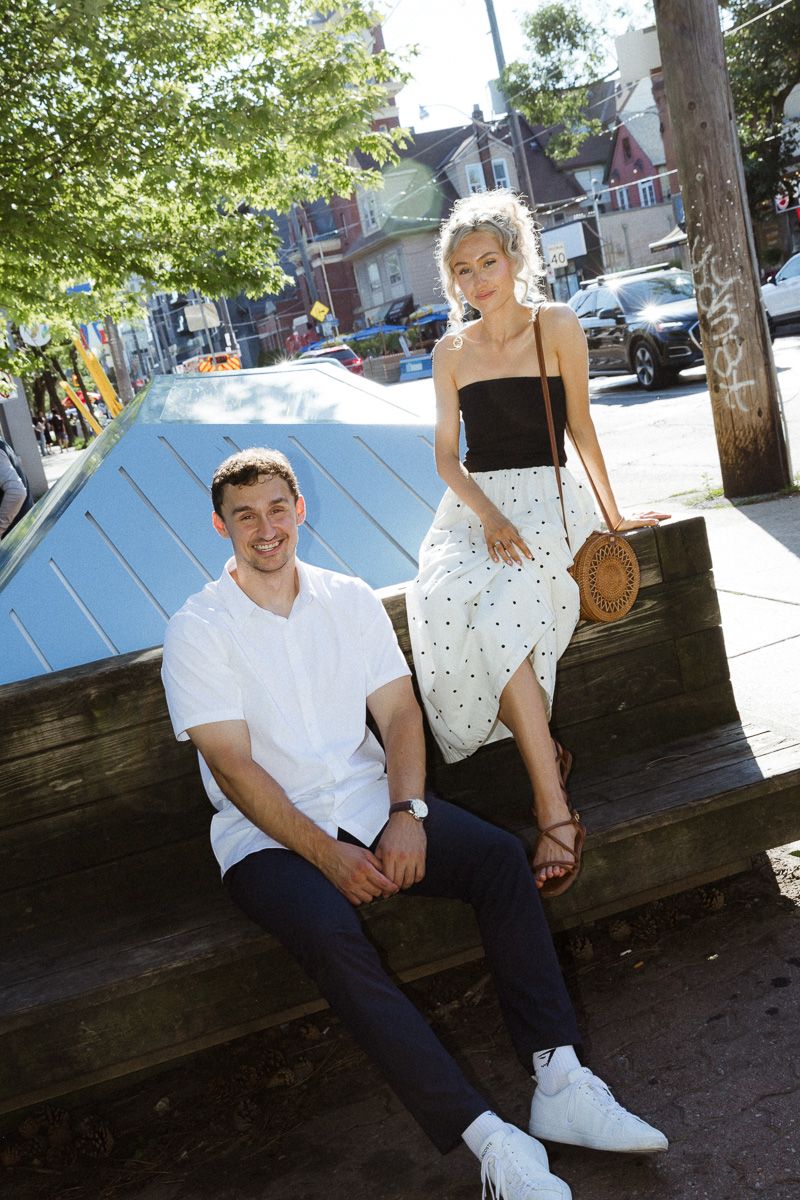 Young couple posing outdoors on a wooden bench in an urban setting, woman in polka dot skirt and man in white shirt.
