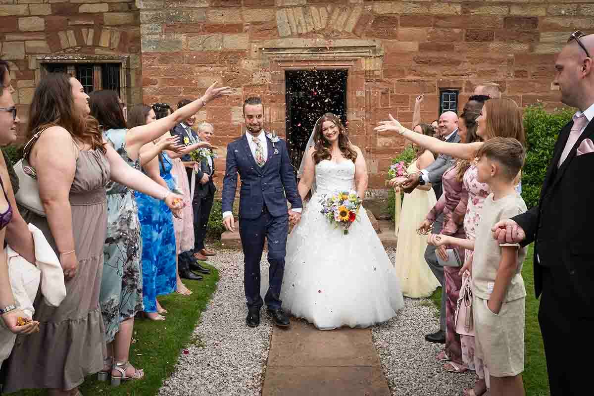 Bride and groom walk through confetti shower with smiling wedding guests lining the path outside Dalston Hall