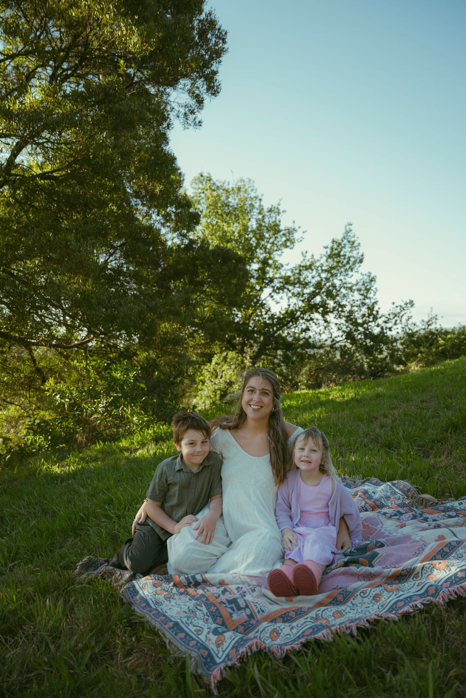 A smiling woman sits on a colorful blanket in a green meadow with two young children beside her.