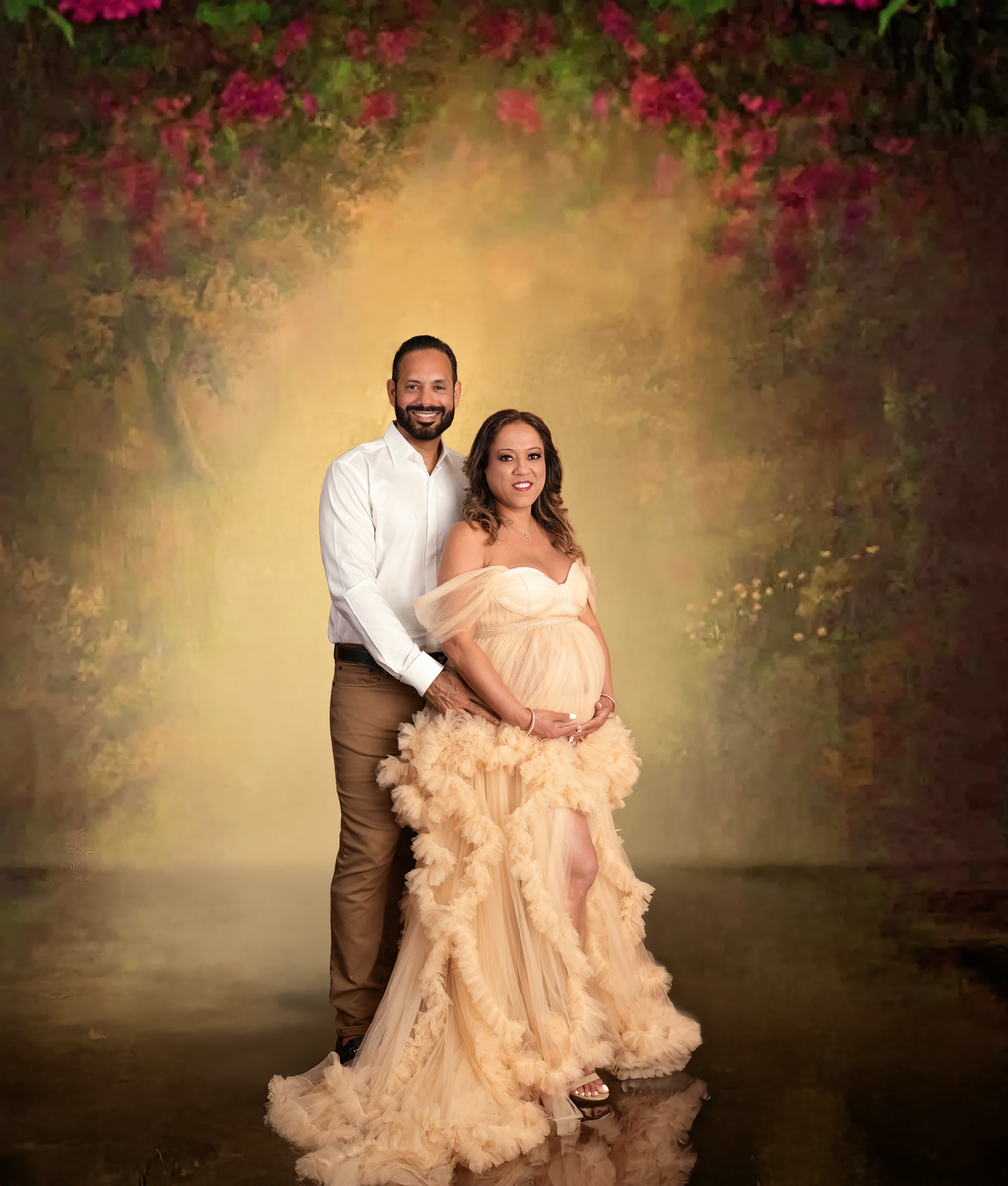 Couple maternity photo shoot with woman in elegant peach ruffle gown and man in white shirt against floral backdrop.