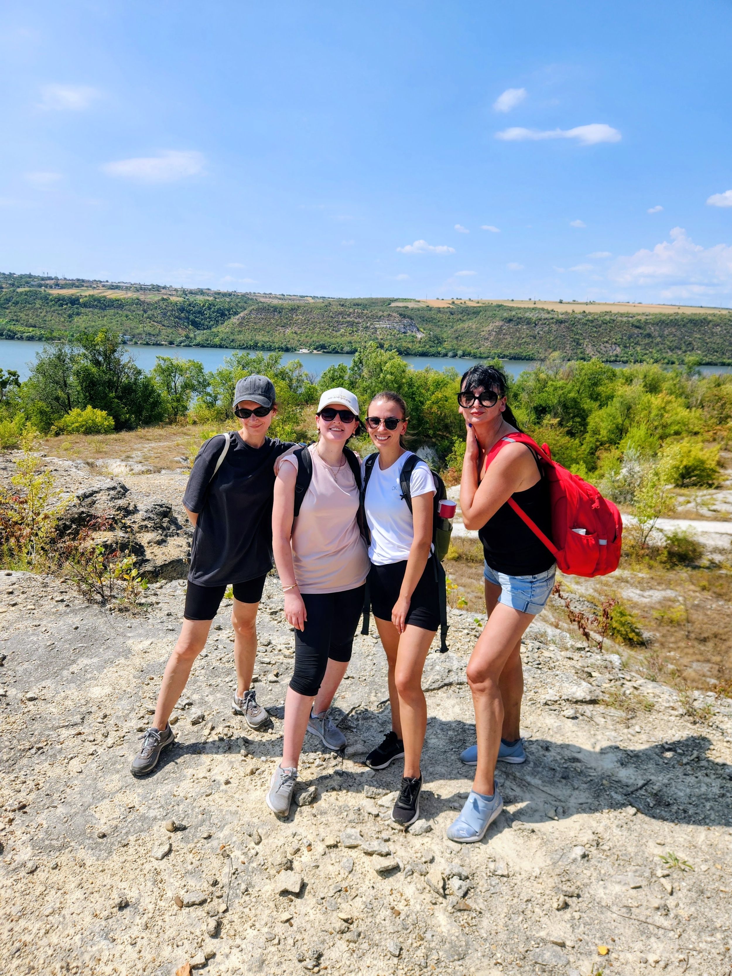 Four women hikers posing on rocky terrain with scenic river valley view on sunny day.