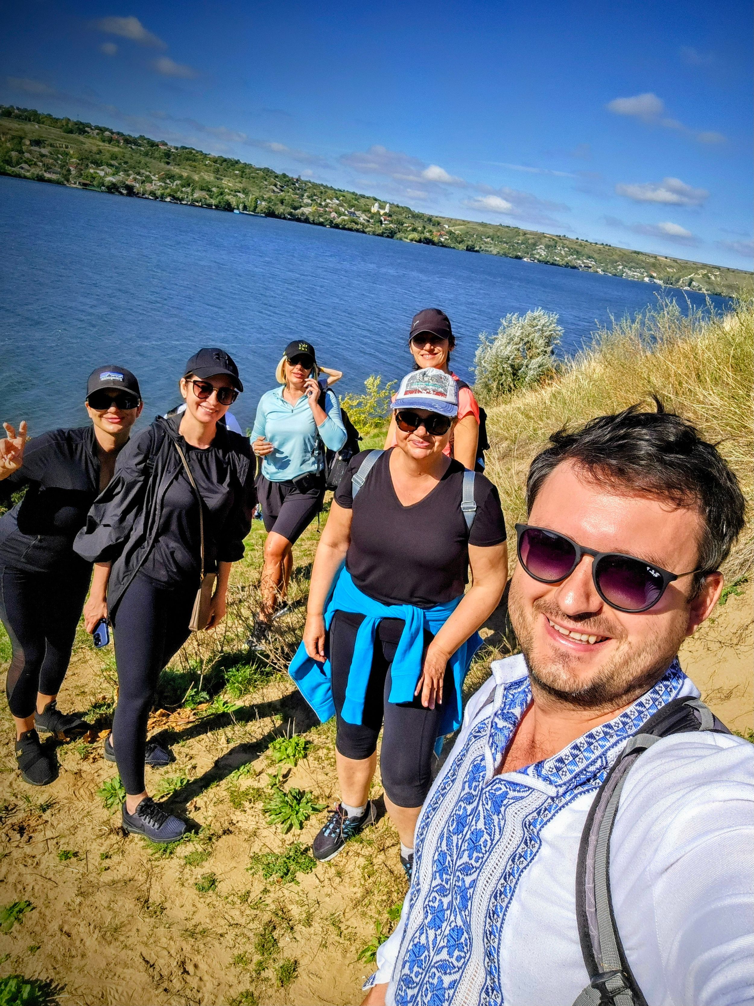 Group of hikers taking a selfie on a sandy cliff overlooking a scenic blue lake on a sunny day.