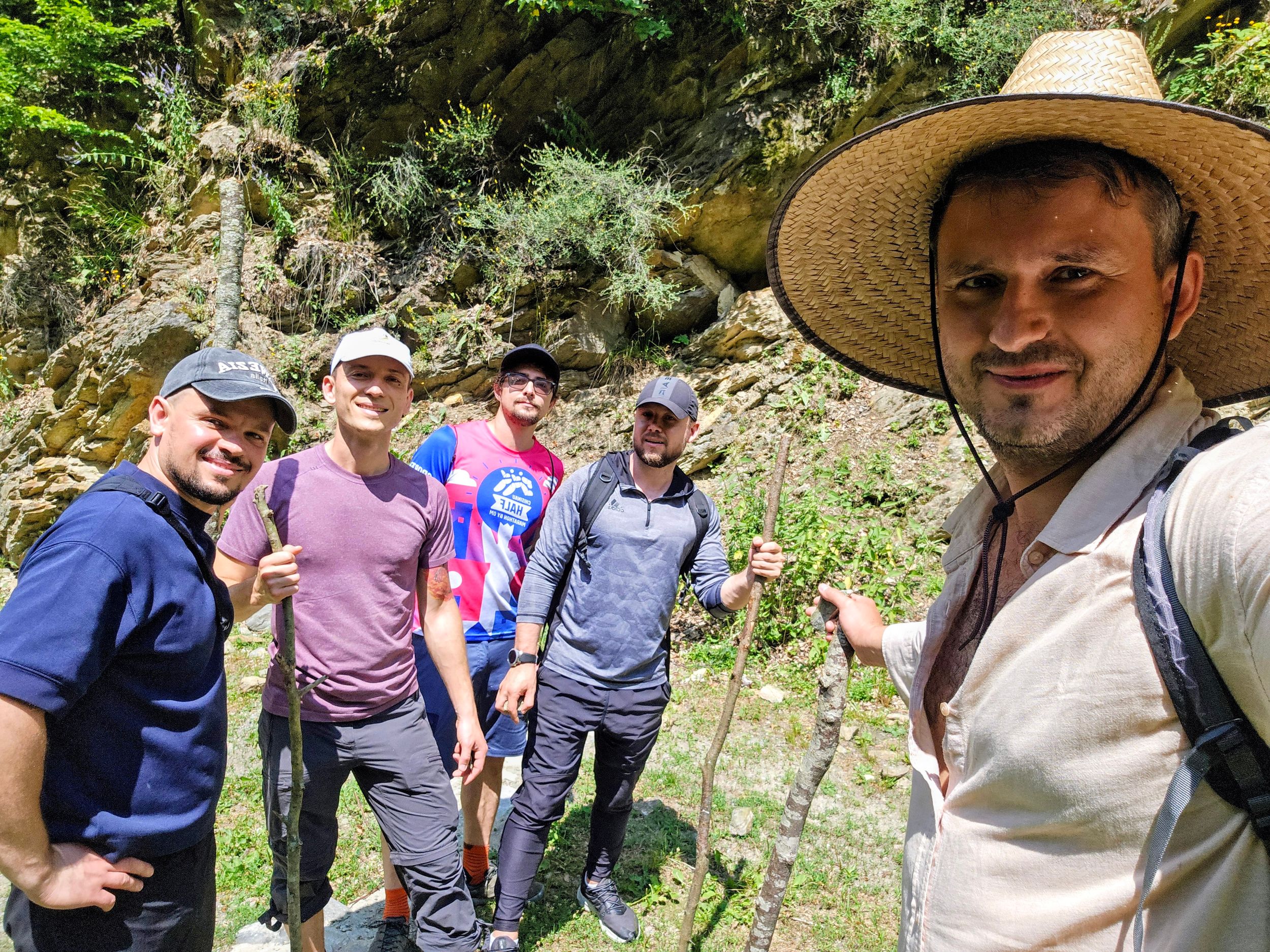 Group of five male hikers taking a selfie in a rocky canyon surrounded by lush green vegetation.