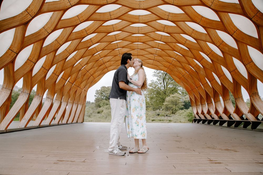 Couple sharing a romantic kiss under a stunning geometric wooden arch structure in an outdoor setting.