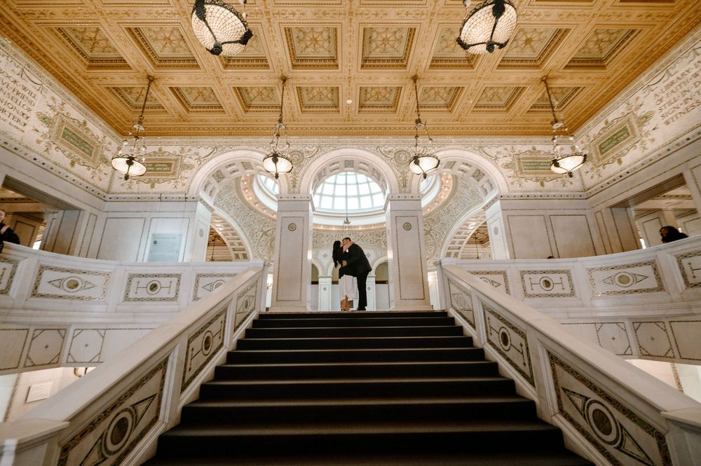 Couple shares romantic moment on grand staircase in elegant white marble hall with ornate coffered ceiling.