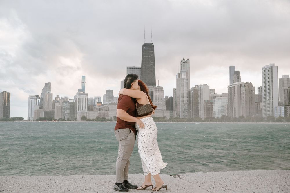Couple embracing on Chicago lakefront with iconic skyline and John Hancock Center in the background on a cloudy day.