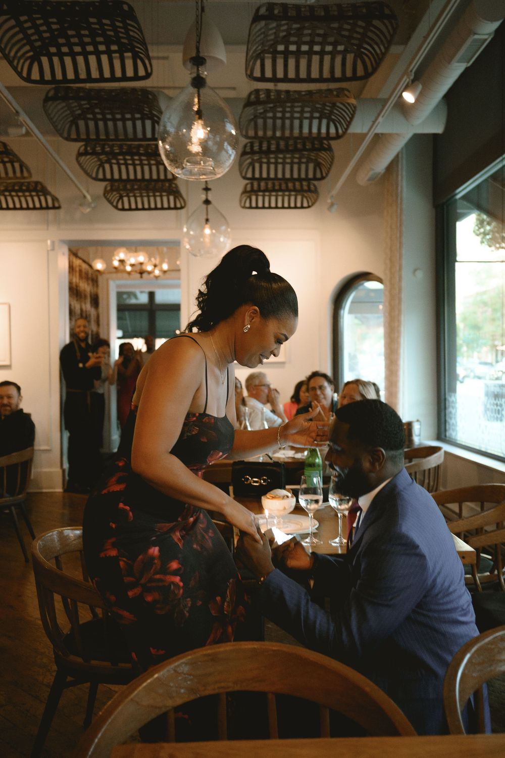 A waitress serves a couple at a candlelit restaurant table, with other diners visible in the background.