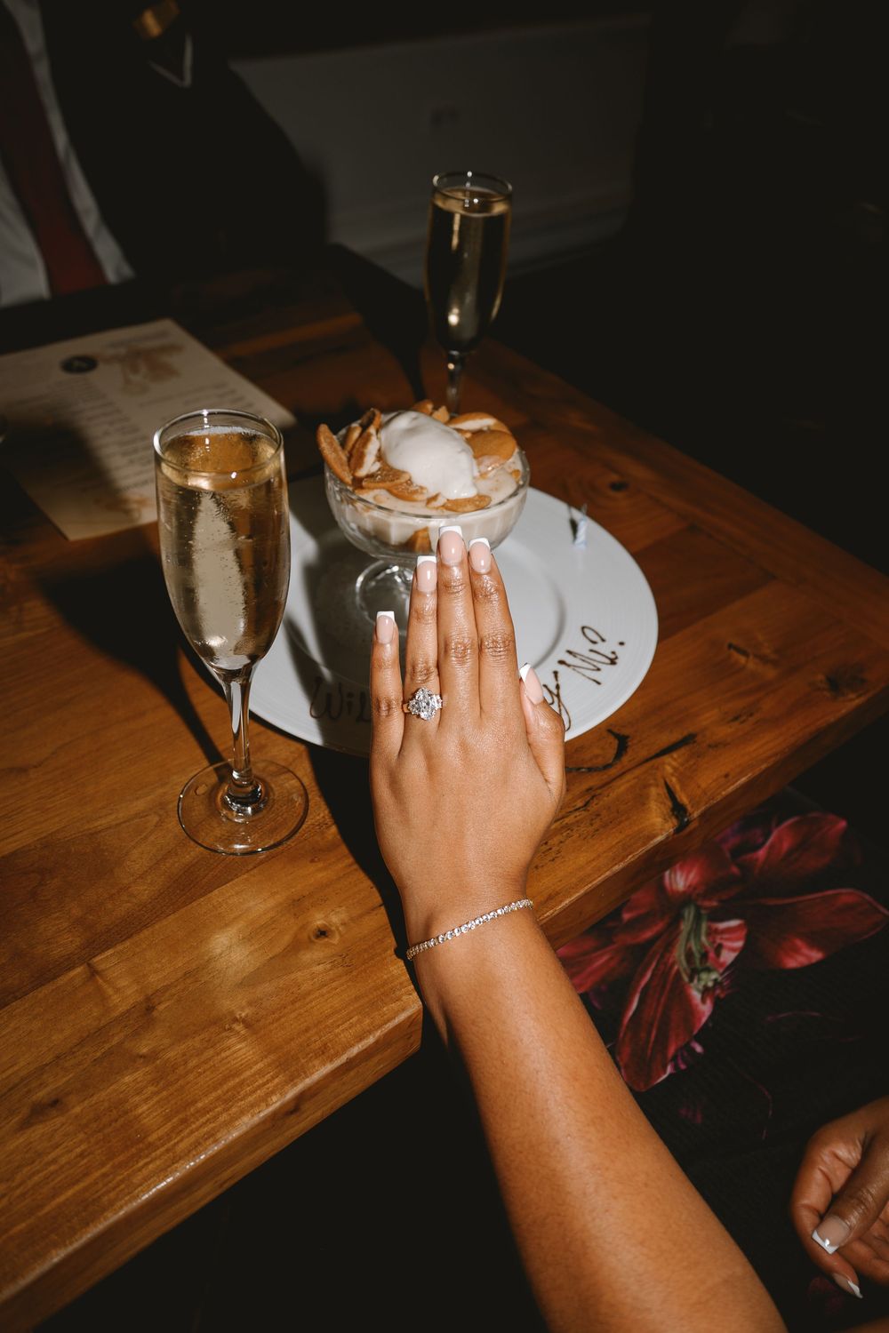 Hand displaying an engagement ring and bracelet near champagne flutes and dessert on a wooden table.