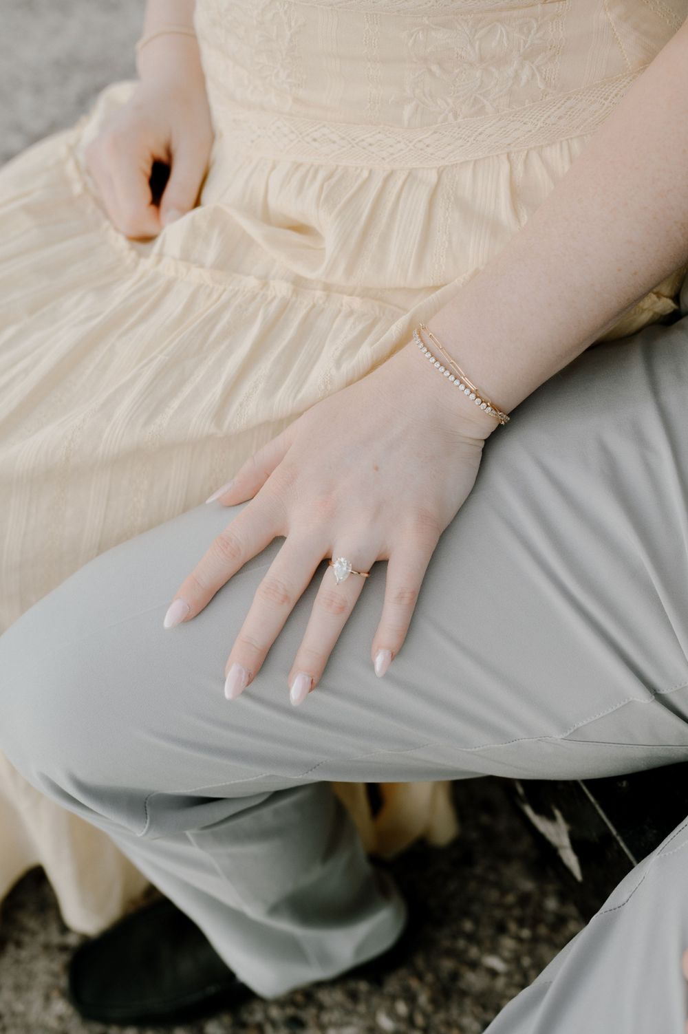 Close-up of a bride's hand displaying an engagement ring and delicate bracelet, resting on gray trousers.