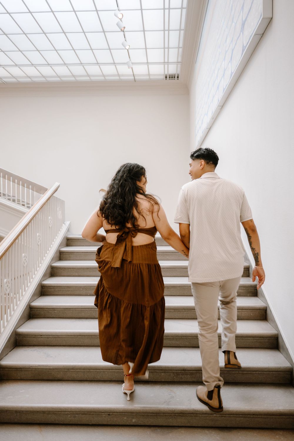 Couple holding hands walking up stairs, woman in brown dress and man in white shirt, viewed from behind.
