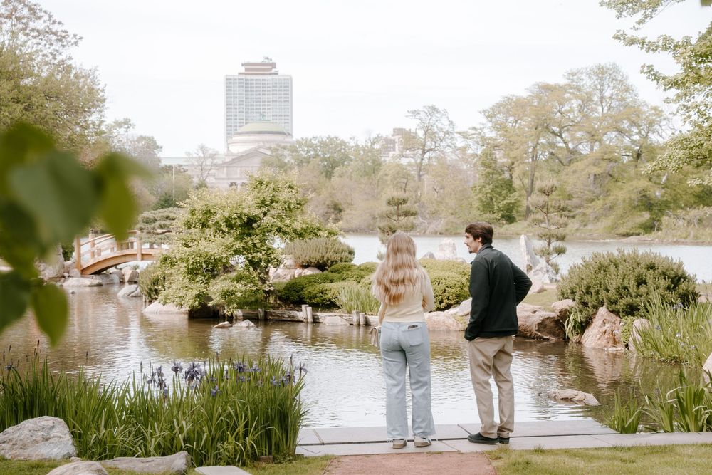 Couple standing hand-in-hand by a serene Japanese garden pond with a red bridge and city skyline in the background.