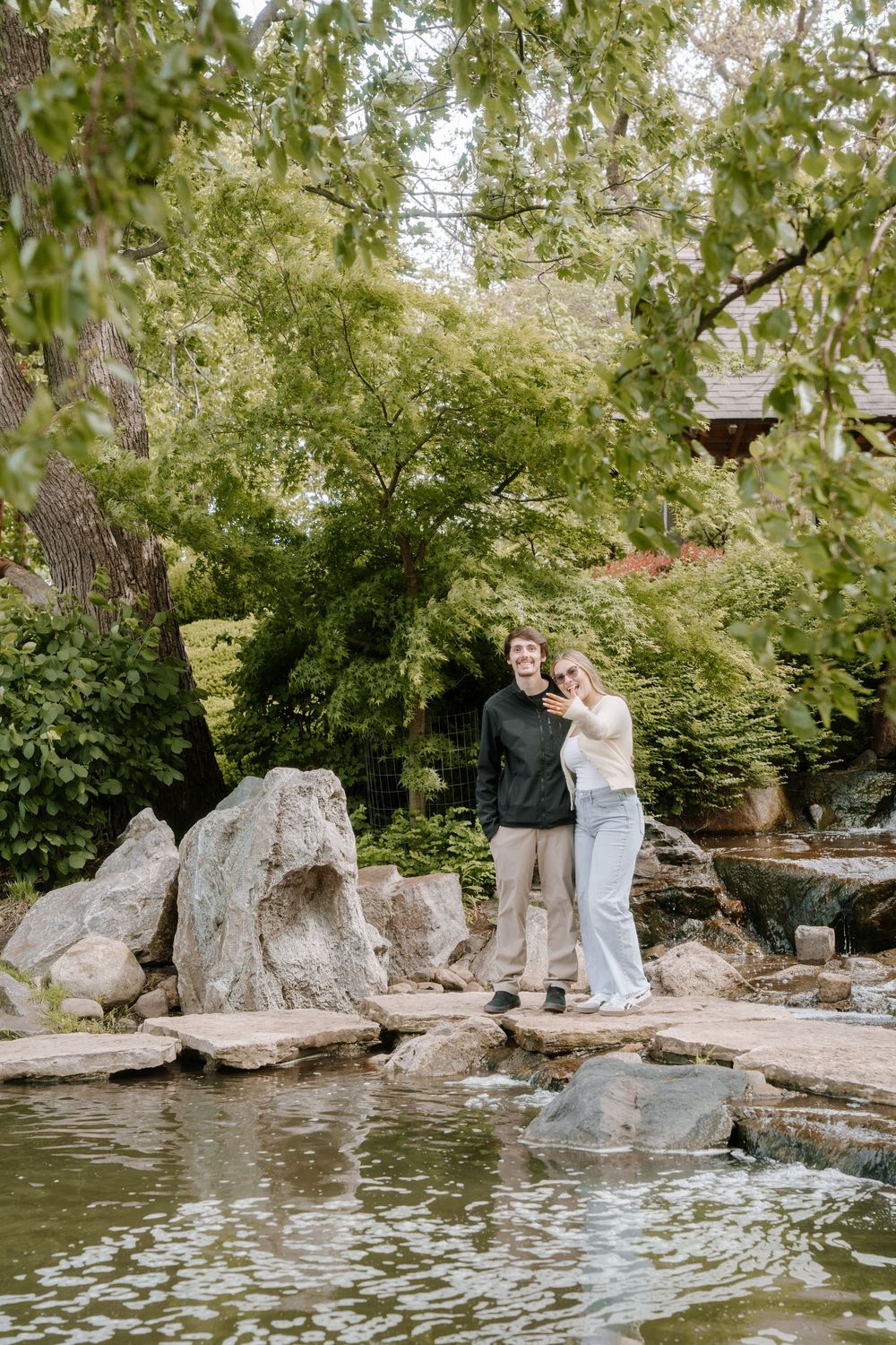 Couple standing on rocks by a pond in a lush green garden, smiling and embracing outdoors.