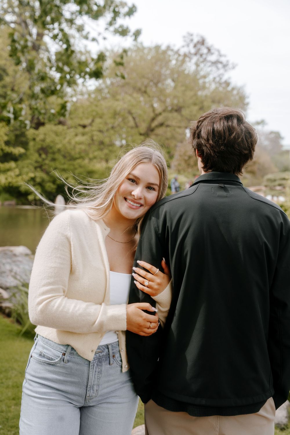 A smiling blonde woman in a cream cardigan and jeans stands with a man in a black jacket in a lush green park.