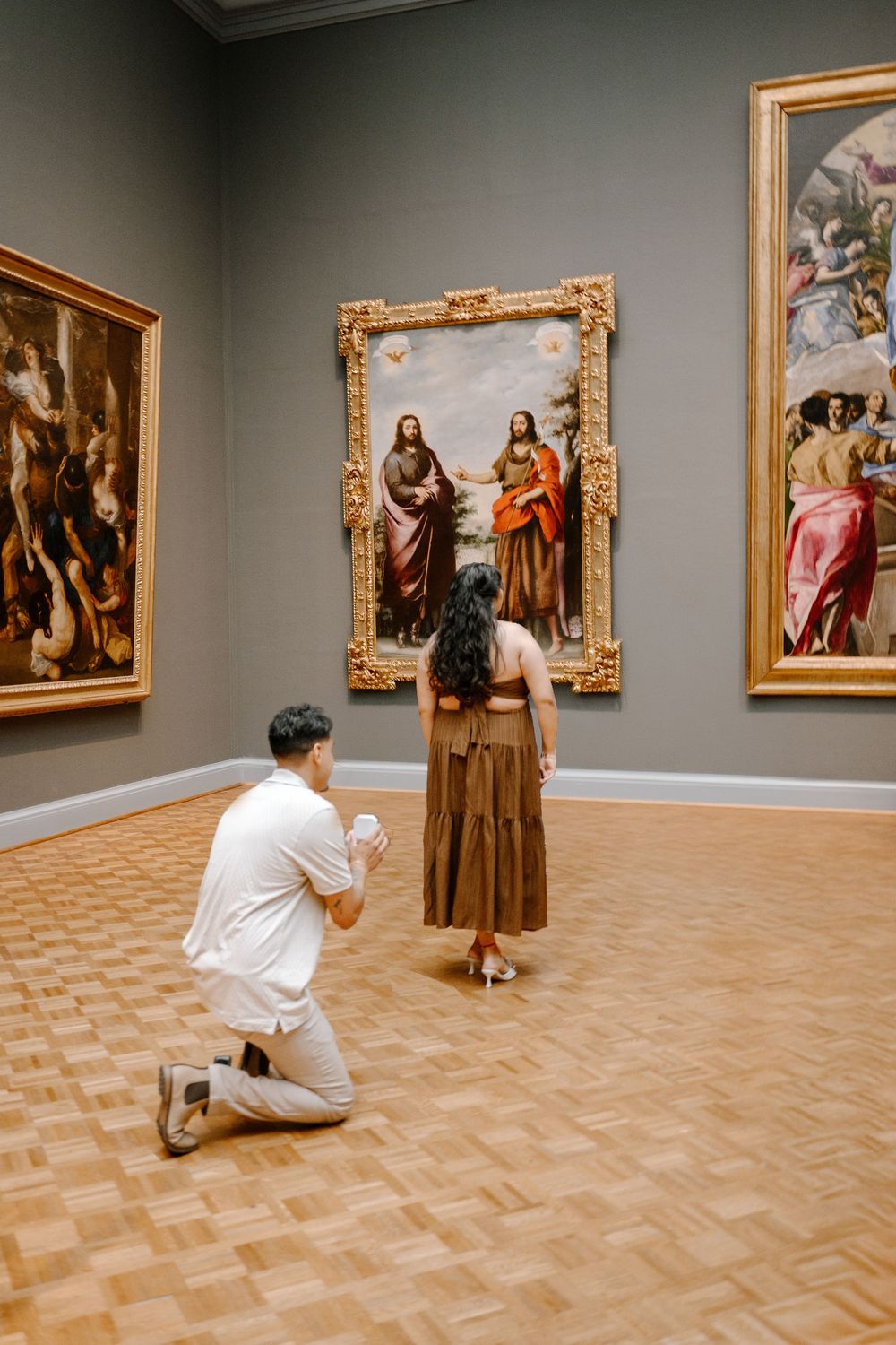 Man proposing on one knee to woman in brown dress at art museum gallery with classical paintings.